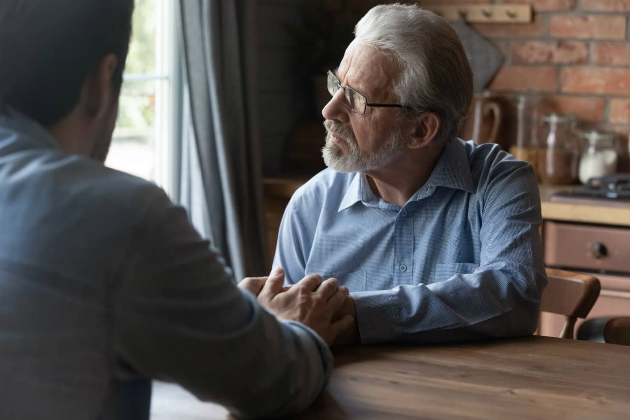 Father stares out of the window sitting at the table with his son making a tough choice on live in care